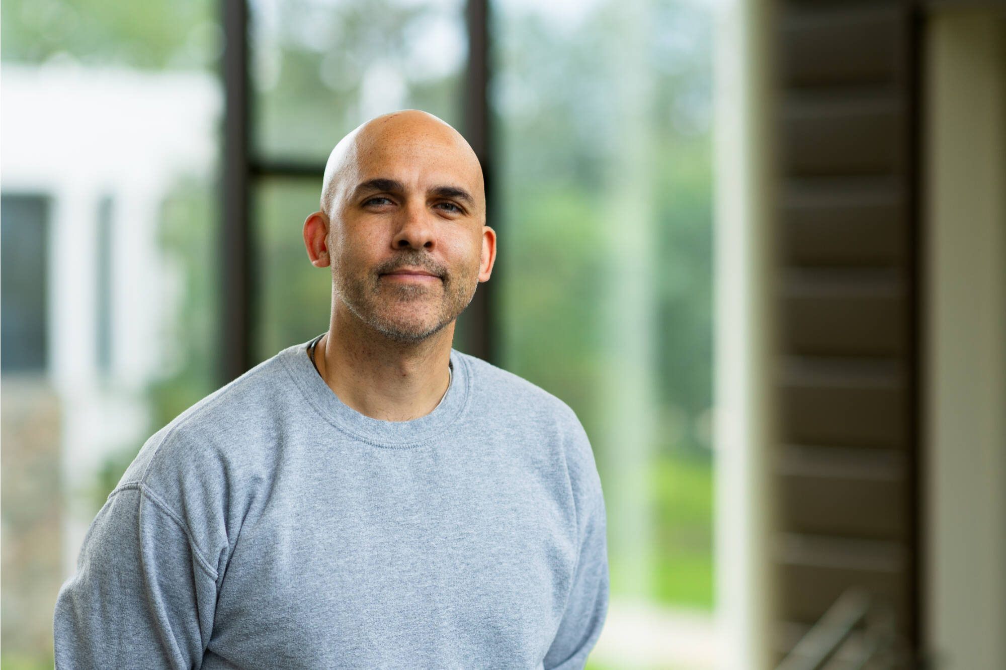 Portrait of a smiling bald man wearing a grey crewneck, standing in front of a blurred green background.